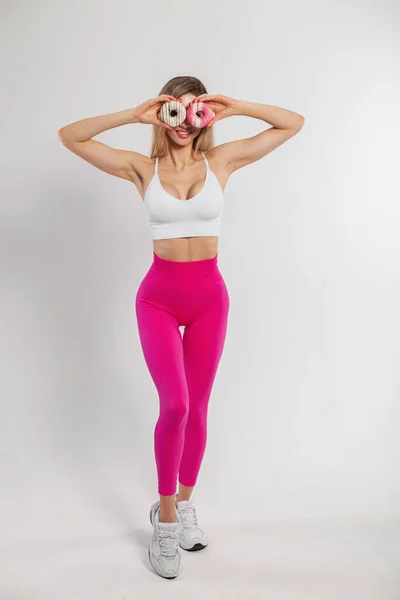 Funny beautiful young fitness lady in fashionable sportswear holds donuts near her eyes and poses on a white background