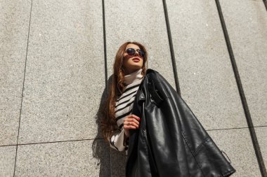 Güneş gözlüklü, şık, siyah deri ceketli ve kazaklı, güneşli bir günde şehrin duvarının yanında duran hippi tarzı güzel bir kız.