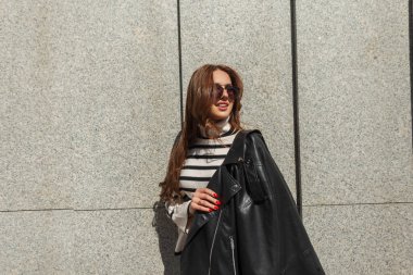 Mutlu, genç, güzel, hippi, gözlüklü, gözlüklü, modaya uygun, süveterli, deri ceketli, şehirde duvarın yanında siyah ceketli, güzel bir kız.