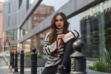 Şık çizgili süveteri olan havalı, genç ve güzel bir kadın model şehirde modern bir alışveriş merkezinin yanında oturuyor.