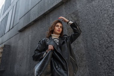 Rock kentsel tarzı giyinmiş, siyah deri ceketli ve çantalı moda şehirli güzel kadın şehirdeki modern siyah bir binanın yanında yürüyor.