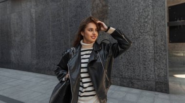 Havalı moda hippisi, güzel kadın model, gülümseyen şehir modasına uygun giyinen, süveterli, deri ceketli ve çanta çantalı siyah modern bir binanın yanında yürüyor.