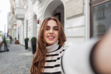 Şık çizgili süveteri olan güzel, güzel ve mutlu bir kadın şehirde yürüyor ve akıllı telefonda selfie portresi yapıyor. Kız Avrupa 'da seyahatin tadını çıkarıyor