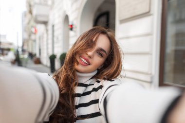 Şık çizgili süveter içinde güzel bir gülüşü olan, Avrupa 'da seyahat eden ve akıllı telefondan selfie çeken sevimli genç bir manken.