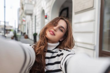 Şık çizgili kazaklı güzel moda genç bir kadın şehirde yürüyor ve akıllı bir telefonla selfie çekiyor. Kadın seyahat eder ve fotoğraf çeker.