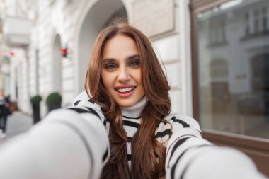 Moda çizgili süveter içinde sevimli gülüşü olan genç, mutlu bir kadın Avrupa şehrinde beyaz bir vintage binanın yakınında seyahat etmekten ve akıllı telefondan selfie çekmekten hoşlanıyor.