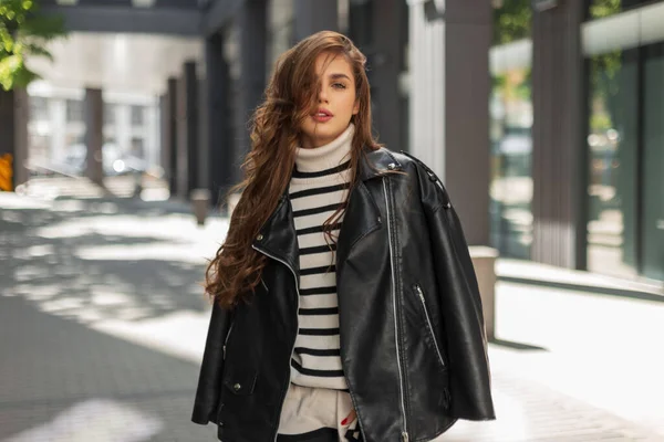 スタイリッシュなレザージャケットと縞模様のセーターを着た流行の服の美しい若いファッショナブルなブルネットの女の子は晴れた日に街を歩く — ストック写真
