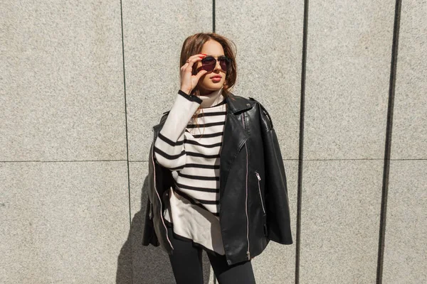 ファッショナブルな美しい女性モデルでスタイリッシュな都会のカジュアル衣装で革黒ジャケットとセーターは 市内の晴れた日に壁の近くのファッションサングラスを身に着けています — ストック写真