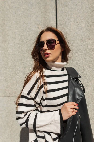 スタイリッシュなサングラスと革のジャケットとストライプのセーターが街の壁の近くに立っているクールな美しいファッション女性モデル太陽の光 — ストック写真