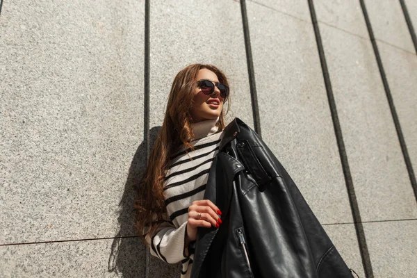 在一个阳光灿烂的城市里 穿着时尚条纹毛衣 穿着时尚皮革黑色夹克 戴着太阳镜的快乐都市时尚女孩站在墙边 街头时尚与美 — 图库照片