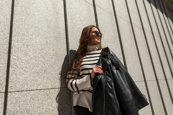 美しい都会的なファッショナブルな女性は 革のジャケットと縞模様のセーターが晴れた日に壁の近くに立っているスタイリッシュな服でサングラス — ストック写真