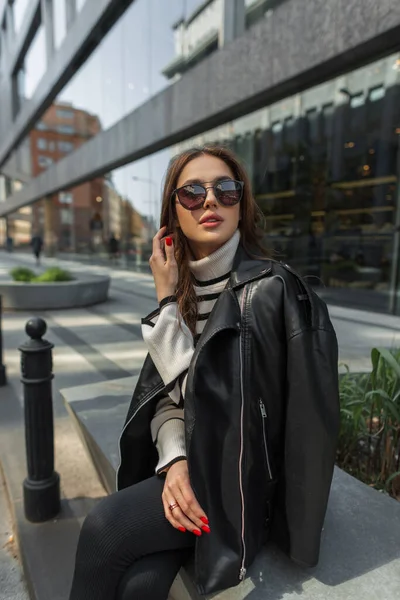 时尚漂亮女孩 身穿休闲装 身穿皮夹克和毛衣 戴着太阳镜 坐在一座现代化建筑附近的城市里 — 图库照片