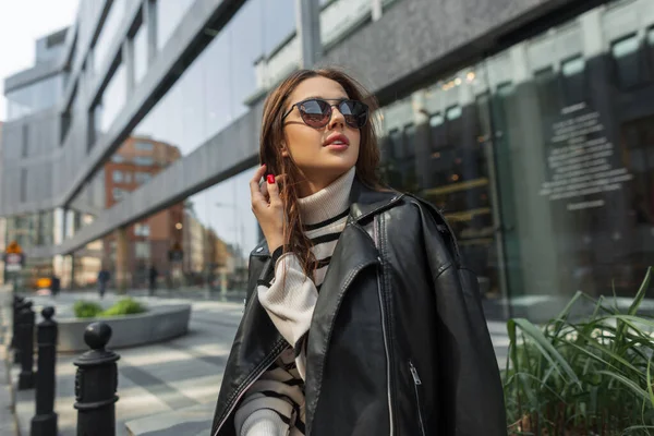 縞模様のセーターと革の黒い岩のジャケットとスタイリッシュな都会の服でサングラスファッション美しい女性モデルは 現代的なモールの近くの街の通りに座っています — ストック写真