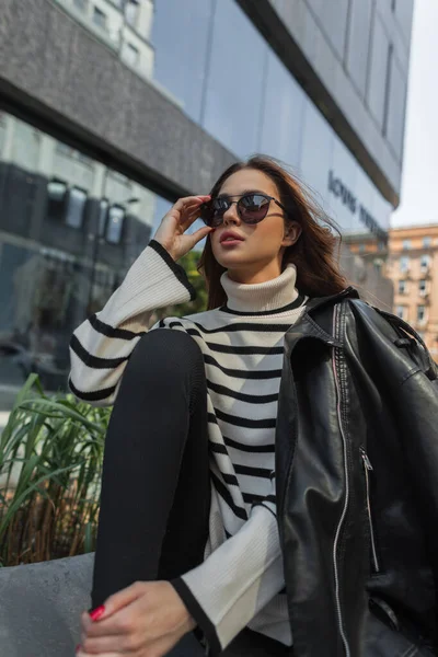 縞模様のセーターと黒の革のジャケットを着たカジュアルな都会の服のファッション美しい女性が座っており 近代的なモールの近くの通りにサングラスをかけて — ストック写真