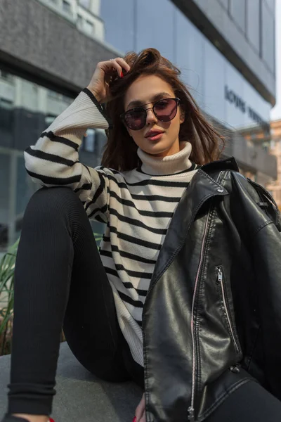 ファッションで流行のサングラスを持つファッショナブルな若い美しい女性セーターと黒の革のジャケットが座っていると通りにポーズ — ストック写真