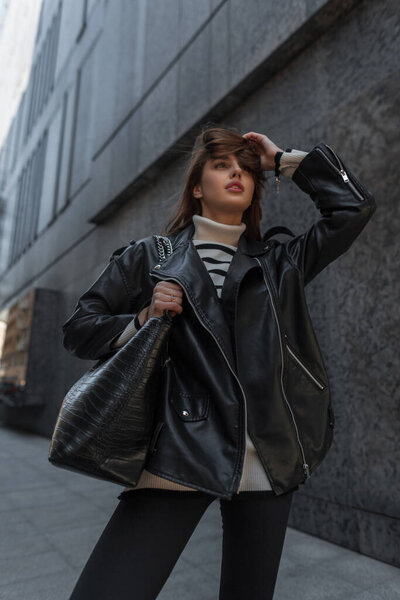 Fashionable young beautiful girl in fashion urban rock outfit with black leather jacket and sweater with bag walks in the city near a black modern building