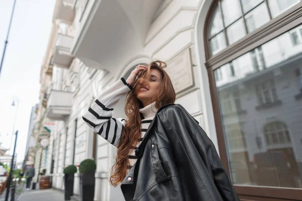 縞模様のセーターと革の黒いジャケットを着たファッショナブルな服の幸せな美しい若い女の子モデルのブルネットは ヴィンテージの白い建物の近くの通りを歩いています ヨーロッパ旅行 — ストック写真