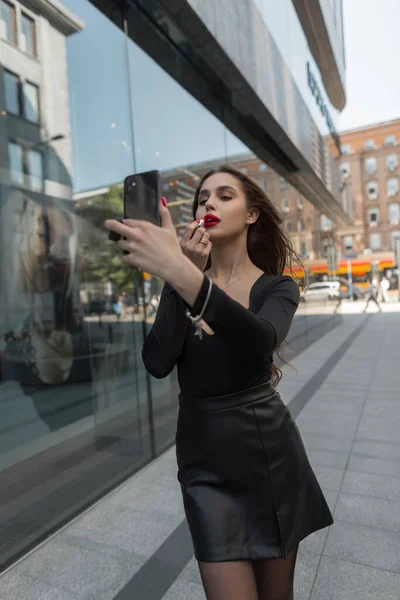 Beautiful young pretty chic woman model paints her lips with red lipstick and looks into a smartphone in the city