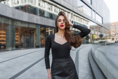 Moda kıyafetleri içinde kırmızı dudakları olan şık, genç bir kadın model şehirde yürüyor ve poz veriyor.