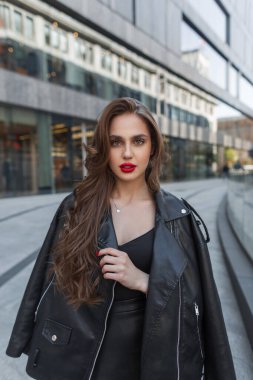 Moda siyah elbiseli, deri ceketli, kırmızı dudaklı çekici bir kız şehirde yürüyor ve kameraya bakıyor.