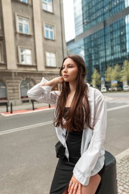 Moda elbiseli, siyah elbiseli ve beyaz gömlekli şık bir kadın mankeni şehirde oturuyor.