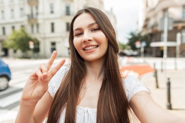 Şık, beyaz elbiseli, mutlu, genç bir kadın şehirde yürüyor, akıllı bir telefonla selfie çekiyor ve barış işareti gösteriyor.