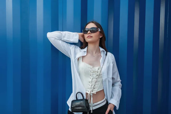 时尚漂亮的少女 戴着时髦的太阳镜 身穿衬衫 紧身衣和手提包 站在蓝色的金属墙边 — 图库照片