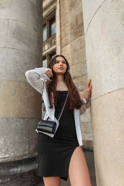 在城市里 穿着白色时髦衬衫和黑色连衣裙 头戴手提包的时髦漂亮女模特站在柱子旁边 — 图库照片