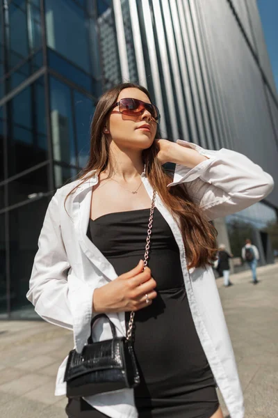 白のシャツとハンドバッグが街を歩くとファッション黒のドレスでサングラスを持つ美しい若いスタイリッシュなエレガントな女の子モデル — ストック写真