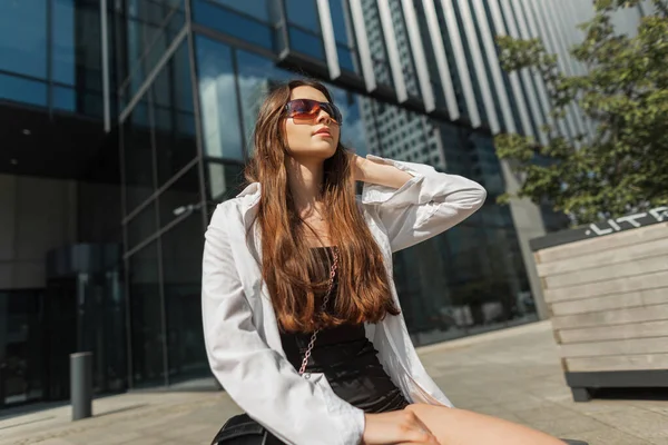 在城市的长椅上坐着一位年轻貌美的时髦女子 戴着太阳镜 穿着时髦的黑色连衣裙 白衬衫 — 图库照片
