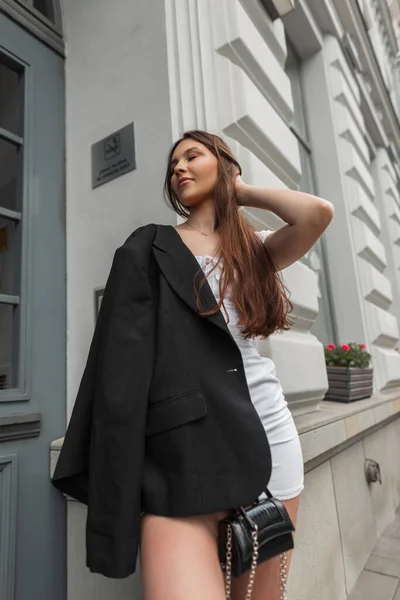 时尚的年轻貌美的女模特站在室外一座老式的白色建筑旁边 她穿着白色衣服 穿着黑色时髦的夹克和手提包 — 图库照片