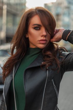 Makyajlı, siyah deri ceketli, yeşil süveterli şık bir bayan şehirde yürüyor ve kameraya bakıyor.