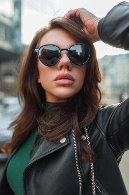 Havalı moda güneş gözlüklü, deri ceketli ve açık havada yeşil kazaklı şık bir kadının şık bir portresi.