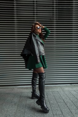 Güneş gözlüklü, havalı, şık, zarif, yeşil elbiseli, açık havada metal duvarın yanında duran siyah deri ceketli, şık, şık, seksi bir kadın.