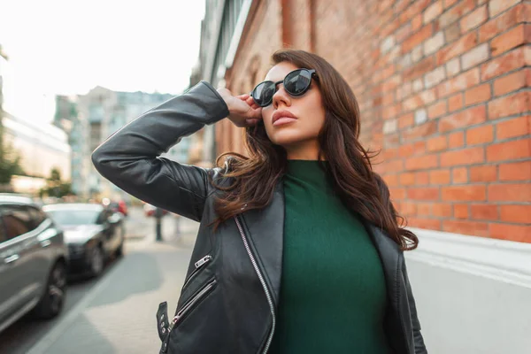 流行の黒革のジャケットと緑のドレスのスタイリッシュな若いトレンディーなシックな女性は 赤レンガの壁の近くに街を歩き サングラスをかける — ストック写真