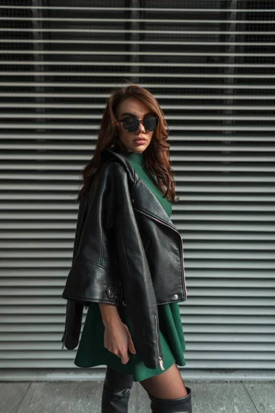 Fashion beautiful woman model with sunglasses in stylish outfit with green dress and black rock leather jacket stands near a metal lines wall in the city