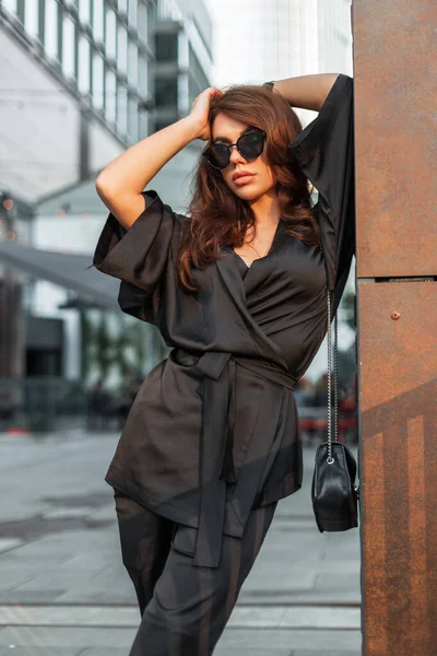 Elegante jovem modelo de mulher urbana bonita com óculos de sol em blazer  de moda preto e top senta-se na rua e posa roupa de estilo de negócios  feminino