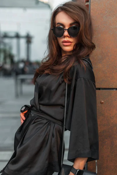 时尚的年轻貌美的女模特 戴着时髦的太阳镜 穿着典雅的黑色西装 手提包 站在城中的一堵墙边 — 图库照片