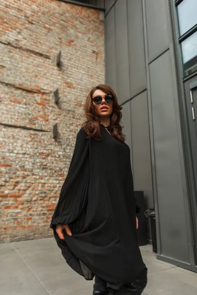 stock image Gorgeous fashion beautiful young lady model with brunette hairstyle with stylish sunglasses in fashionable black dress walks and poses in the city near the building. Pretty glamour woman in outfit