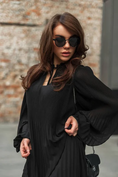 在城中靠近砖楼的地方 一位年轻貌美的女模特 戴着老式太阳镜 身穿时尚时髦的黑色连衣裙 手里拿着一个小手提包 — 图库照片