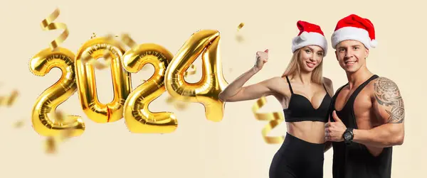Healthy happy athletic couple strong man and slim woman with red santa hat showing muscles and like on beige background happy new year 2024 golden balloons. Health, sport and happy new year