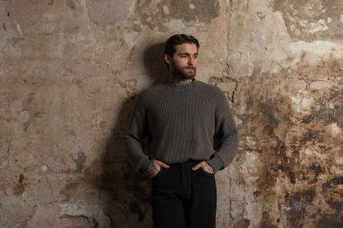 Sakallı, saç stili, desenli süveter ve beton bir duvarın yanında siyah kot pantolon giyen yakışıklı, vahşi bir adam.