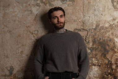 Moda örgü süveteri içinde bıyıklı, sakallı ve saç stili yakışıklı hippi genç adam grunge beton bir duvarın yanında duruyor.