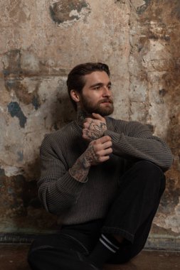 Moda örgü süveter ve siyah kot pantolon dövmeli, saç stili ve sakallı yakışıklı, vahşi hippi stüdyodaki eski bir beton duvarın yanında oturuyor.