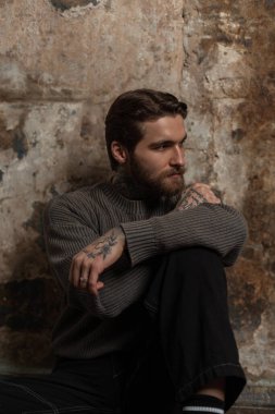 Havalı, sakallı, modaya uygun örgü süveter giyen yakışıklı adam eski moda bir grunge arka planının yanında oturuyor.