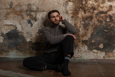 Süveter ve çoraplı siyah kot giyen havalı hippi stüdyodaki eski grunge beton duvarın yanında oturuyor.