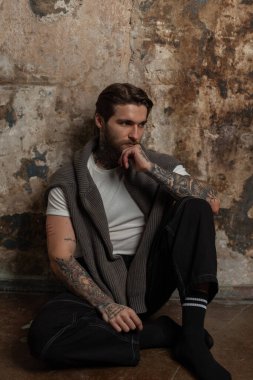 Havalı, havalı, hippi, saç stili ve sakallı beyaz tişörtlü, kazaklı, kot pantolonlu bir adam grunge duvarın yanında oturuyor.