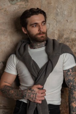 Sakallı, bıyıklı, moda kıyafetli, süveterli ve eski beton duvarın yanında beyaz tişörtlü havalı bir genç adam modeli.