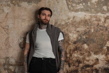 Sakallı, beyaz tişörtlü, örgü kazaklı, saç stili, yakışıklı ve yakışıklı bir adam beton bir duvarın yanında duruyor.