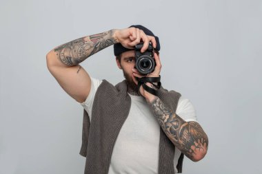 Dövmeli, örgü şapkalı, kazaklı, aynasız fotoğraf makinesi tutan ve stüdyoda fotoğraf çeken yakışıklı bir profesyonel.